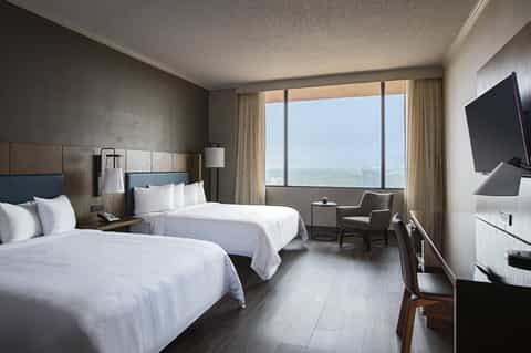 Hotel bedroom with two beds, desk area, and floor-to-ceiling windows with landscape view