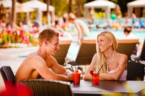 Man and woman enjoying drinks poolside at resort with lounge chairs and tropical landscaping in background
