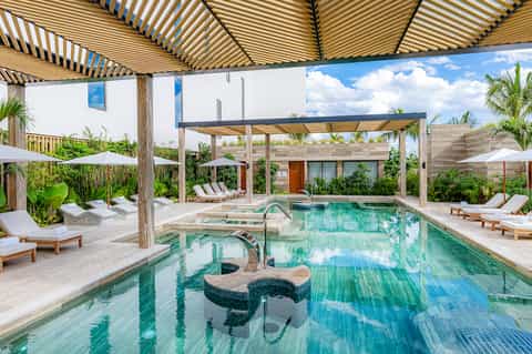 Indoor pool with lounge chairs, wooden pergolas, striped shade structures, and lush tropical plants