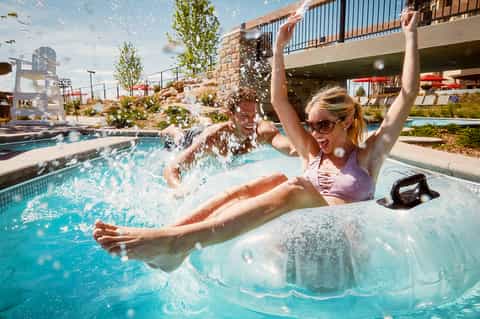 Woman enjoying water feature at resort pool with splash, lounge chairs, and modern architecture visible