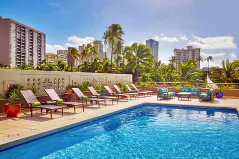 Resort pool deck with lounge chairs, palm trees, and city skyline in background