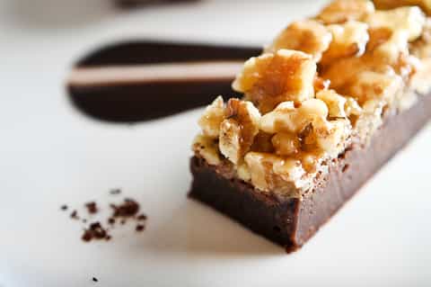 Decadent chocolate brownie dessert topped with caramel and walnuts on white plate