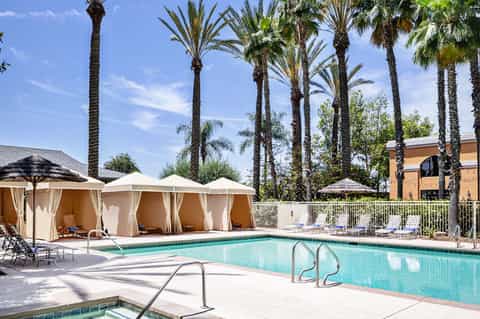 Resort pool with turquoise water, white cabanas with curtains, palm trees, and lounge chairs on sunny day