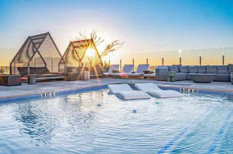 Modern rooftop pool with loungers, geometric shade structures, seating areas, and sunset skyline views