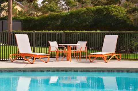 Poolside loungers with orange frames and white cushions beside turquoise water and green hedge privacy fence