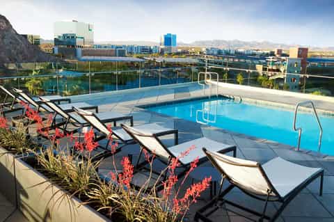 Rooftop pool overlooking urban waterfront with mountains, lounge chairs with white cushions, red flowering plants, and city skyline