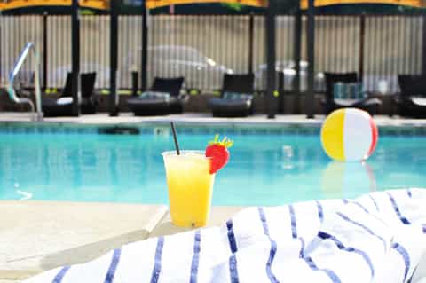 Yellow cocktail with strawberry garnish on pool deck beside striped towel and beach ball
