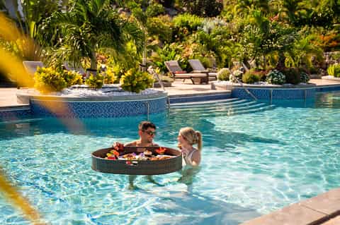 Couple enjoying floating food basket in resort pool surrounded by tropical landscaping and loungers