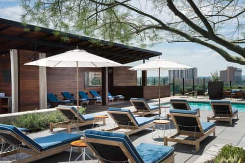 Rooftop pool deck with blue loungers, white umbrellas, wood cabana, and city skyline in background