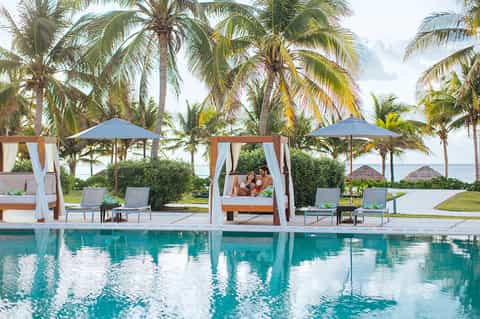 Luxury resort pool with wooden cabanas, blue umbrellas, palm trees, and tropical beachfront setting