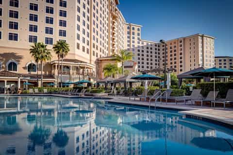 Resort pool with multi-story hotel buildings, blue umbrellas, and palm tree landscaping