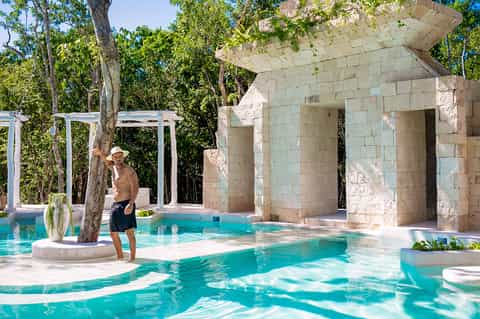 Man standing by turquoise pool with stone structure and lush greenery at luxury resort