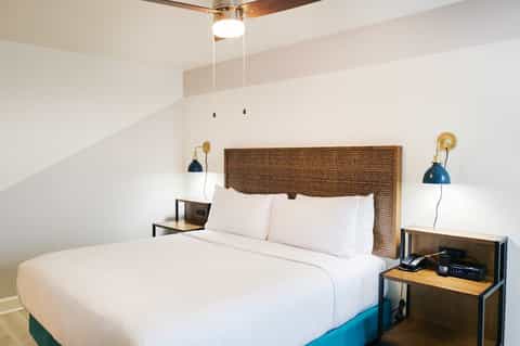 Modern hotel bedroom with woven headboard, white bedding, blue accent bed base, and brass wall sconces
