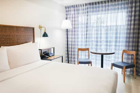Modern hotel bedroom with woven headboard, white bedding, desk lamp, and blue checkered curtains with seating area