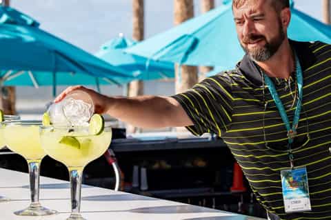 Bartender in striped shirt pouring margarita cocktails poolside under turquoise umbrellas