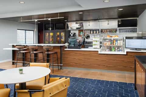 Modern hotel bistro with wood counters, bar seating, white shelving, and casual dining tables with yellow chairs