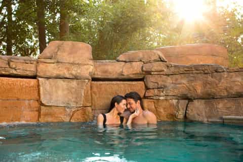 Couple relaxing in turquoise hot spring pool surrounded by rustic stone walls and forest trees