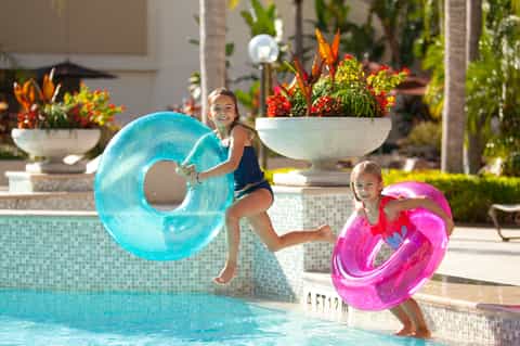 Two children holding colorful inflatable rings by a sunny resort pool with tropical flowers in background