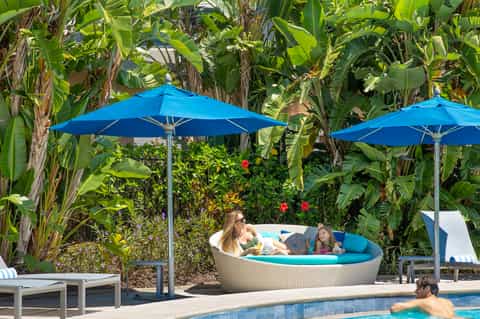 Resort pool area with blue umbrellas, lounge chairs, and lush tropical banana plants in background