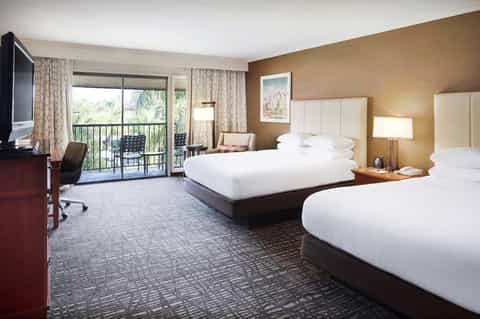 Hotel guest room with two queen beds, balcony access, flat-screen TV, and warm tan walls