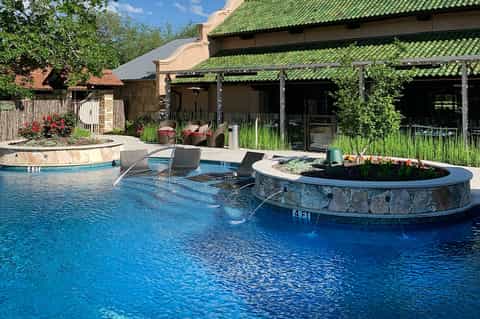 Resort pool with circular fire pit island, lounge chairs, and green-roofed building with pergola