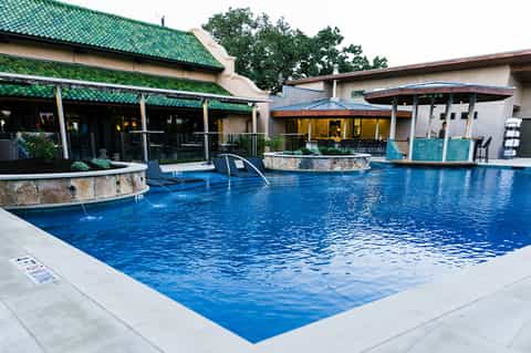 Resort pool with blue water, lounge chairs, green-roofed pavilion, and stone water features surrounded by buildings
