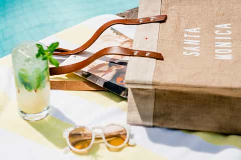 Poolside flat lay with mojito cocktail, sunglasses, beach bag, and leather sandals on white towel