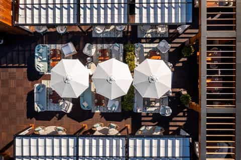 Aerial view of rooftop terrace with three white umbrellas, lounge seating, and dining tables on wooden deck