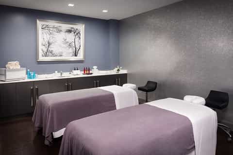 Luxury spa treatment room with two massage beds, purple accent wall, dark cabinetry, and framed botanical artwork