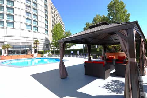 Resort pool area with covered lounge pavilion, black wicker seating with red cushions, and multi-story hotel building