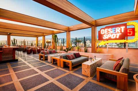 Modern rooftop lounge with wooden beams, orange seating, cacti, and city skyline views with Desert Spot neon sign