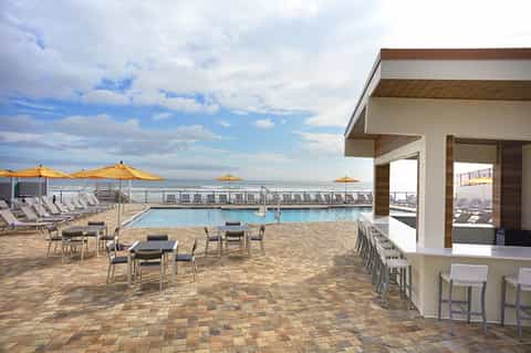 Oceanfront pool deck with yellow umbrellas, lounge chairs, and modern bar pavilion overlooking the sea
