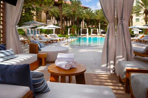 Luxury resort pool area with palm trees, lounge chairs, cabanas with gray curtains, and turquoise water