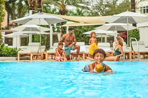 Family enjoying pool with child swimming with yellow ball while adults relax under umbrellas