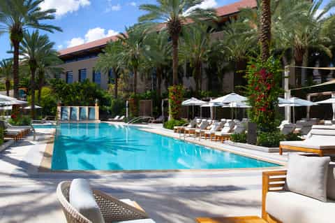 Luxury resort pool with palm trees, lounge chairs, white umbrellas, and Mediterranean-style building