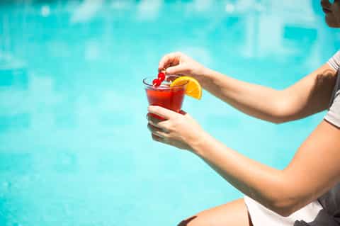 Person holding a red cocktail with orange slice garnish by a bright turquoise swimming pool