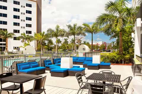 Resort poolside lounge area with blue cushioned seating, palm trees, and white building in background