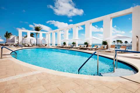 Luxurious resort pool with crystal-clear water, white pergolas, lounge chairs, and ocean views under blue sky