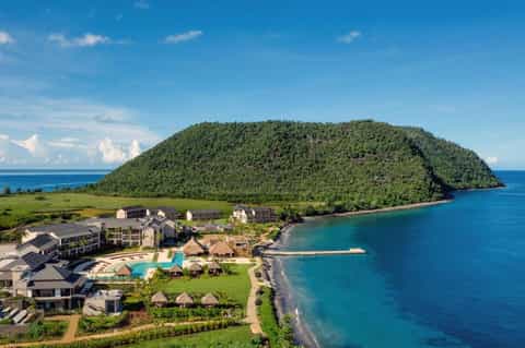 Luxury beachfront resort with pool, thatched bungalows, and forested mountain backdrop overlooking turquoise ocean