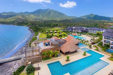 Beachfront tropical resort with multiple pools, thatched-roof structures, and forested mountains in background