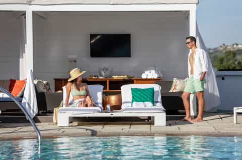 Couple relaxing by an outdoor pool with modern white cabana and entertainment area overlooking hillside