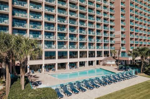 Large beachfront resort with red brick building, balconies, and outdoor pool with lounge chairs