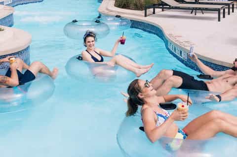 Group of guests floating on inflatable tubes in a bright blue resort pool with tropical landscaping