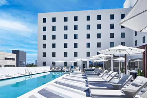 Modern white hotel building with rooftop pool, striped loungers, and white umbrellas under blue sky