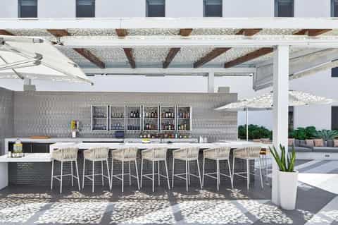 Modern outdoor bar under pergola with white stools, patterned backsplash, and lounge seating in background
