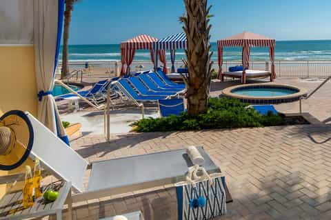 Beach resort patio with blue lounge chairs, striped cabanas, palm tree, and ocean view