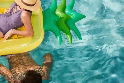 Woman on yellow float raft in turquoise pool water wearing sun hat with green inflatable flamingos