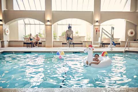 Indoor swimming pool with children playing on inflatable unicorn float, arched windows above