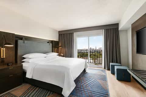 Modern hotel bedroom with dark wood furnishings, white bedding, and floor-to-ceiling windows overlooking a city skyline