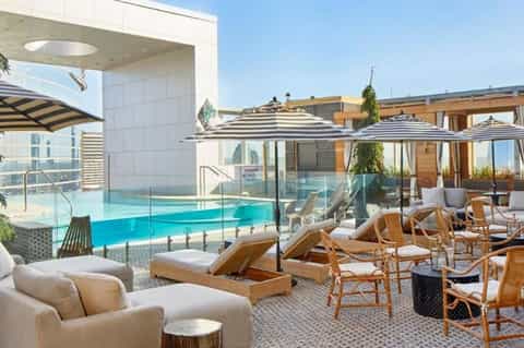 Luxury rooftop pool deck with striped umbrellas, wooden loungers, glass safety barriers, and city skyline views
